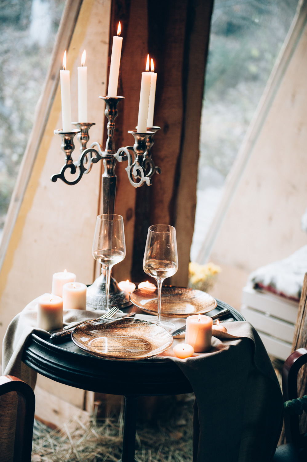 On a small round table are plates and glasses of wine, a large candle holder with candles and several small white candles.