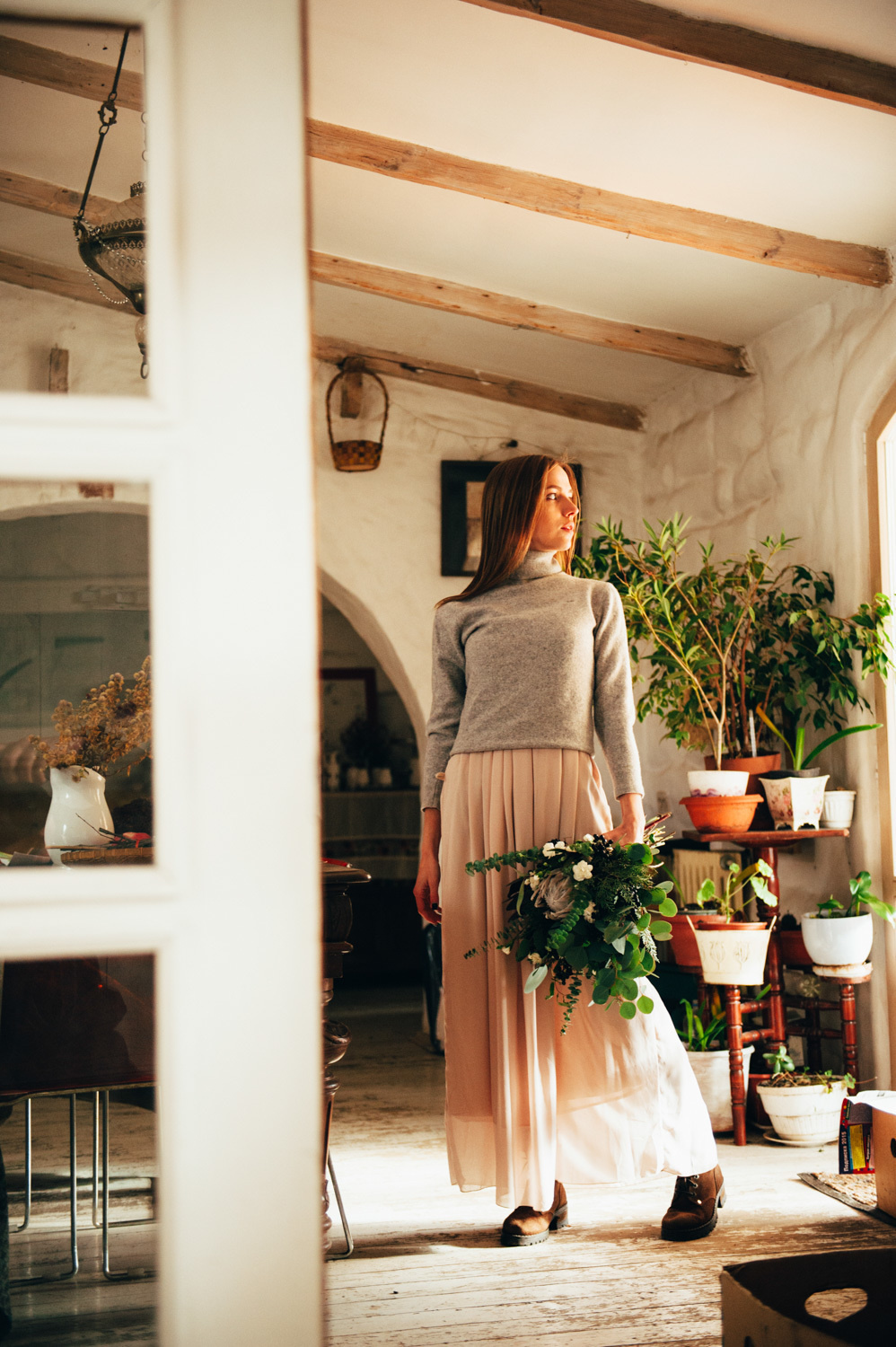 A bride with a bouquet of flowers stands in a room of a village house against the background of a white arch next to pots of indoor flowers.