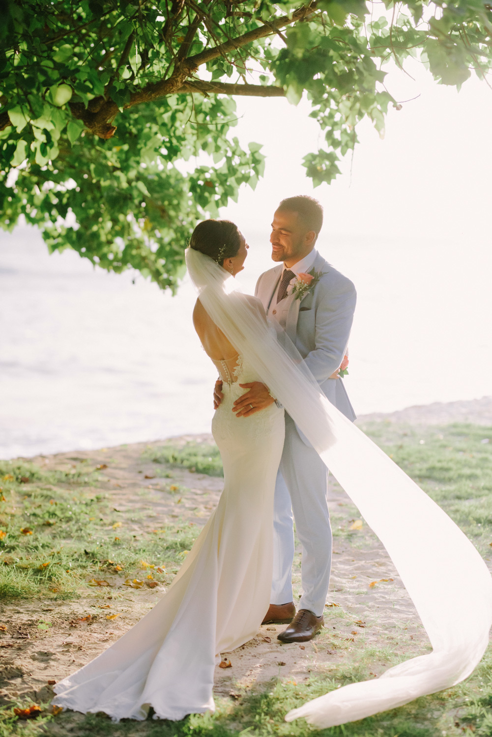 The bride and groom, embracing, stand under a green tree against the background of the sea. The veil flutters in the wind.