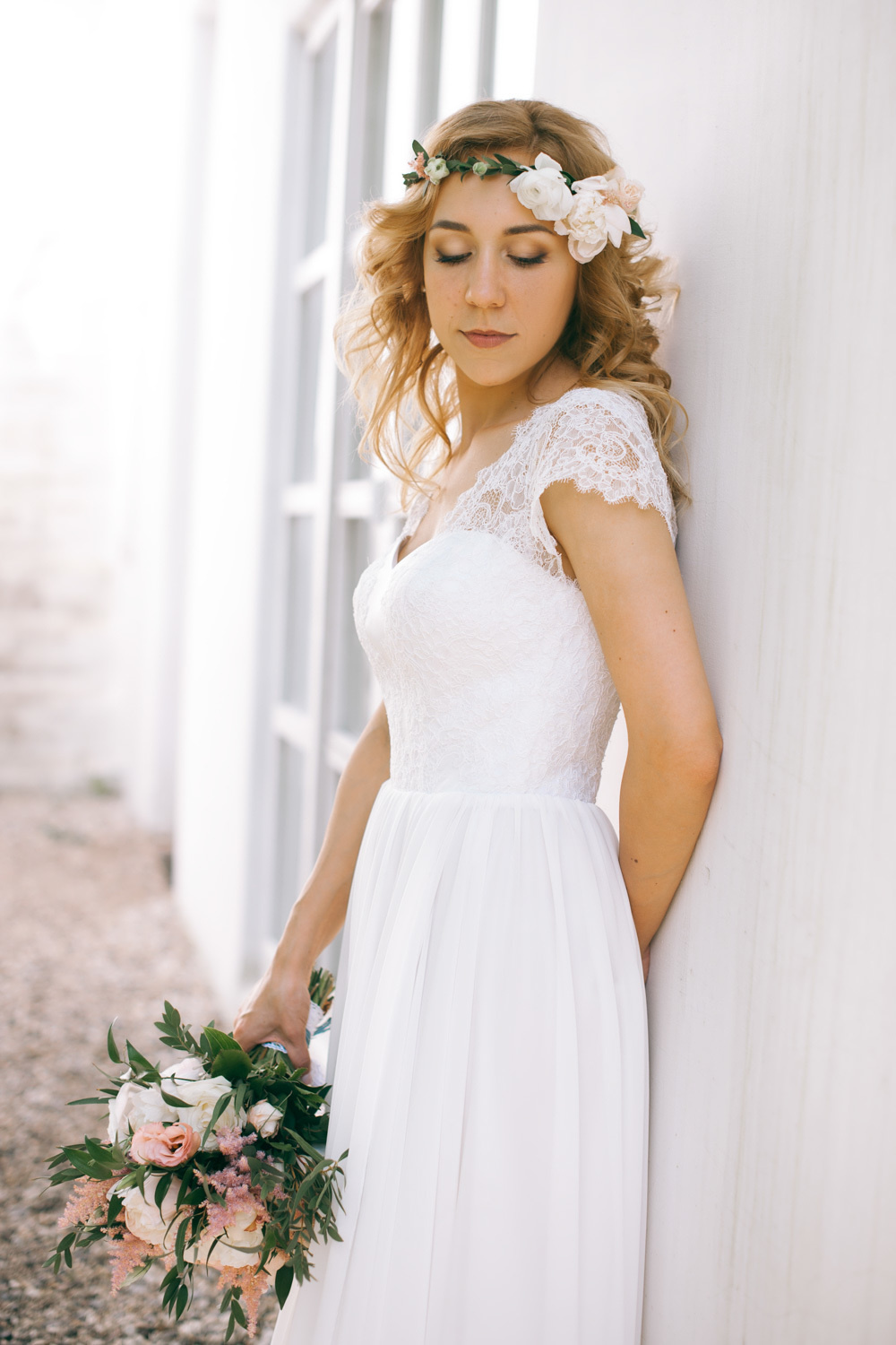 The bride with a bouquet of flowers stands at the white wall.