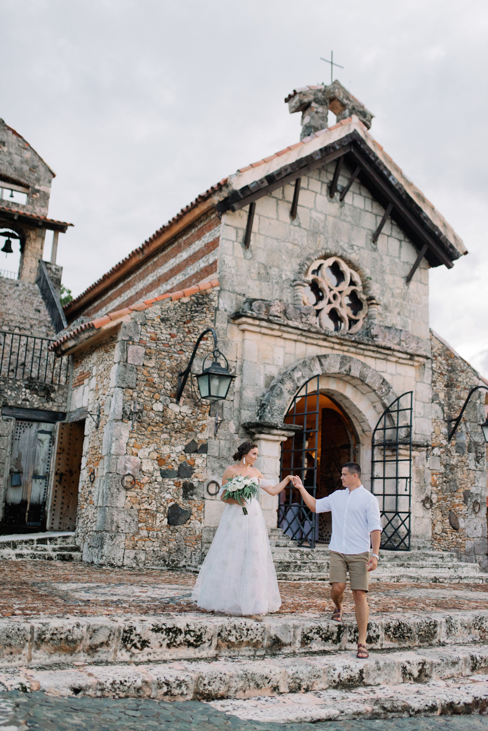 Жених держит невесту за руку на каменных ступеньках перед старинным каменным храмом.