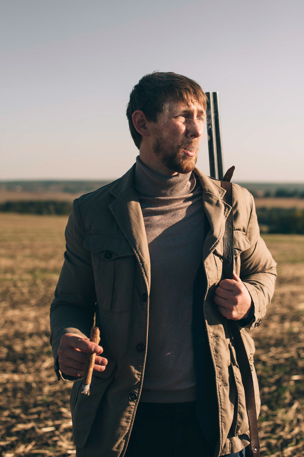 A hunter holds a rifle on his shoulder and a decoy for an animal in his hand against the background of a field and forest.