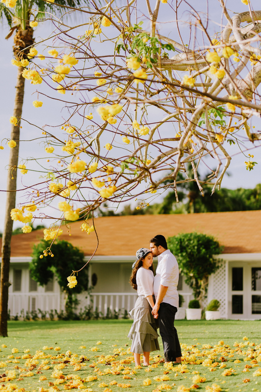 Under a tree with large yellow flowers, the bride and groom stand hand in hand against the background of a one-story white wooden house with an orange roof.