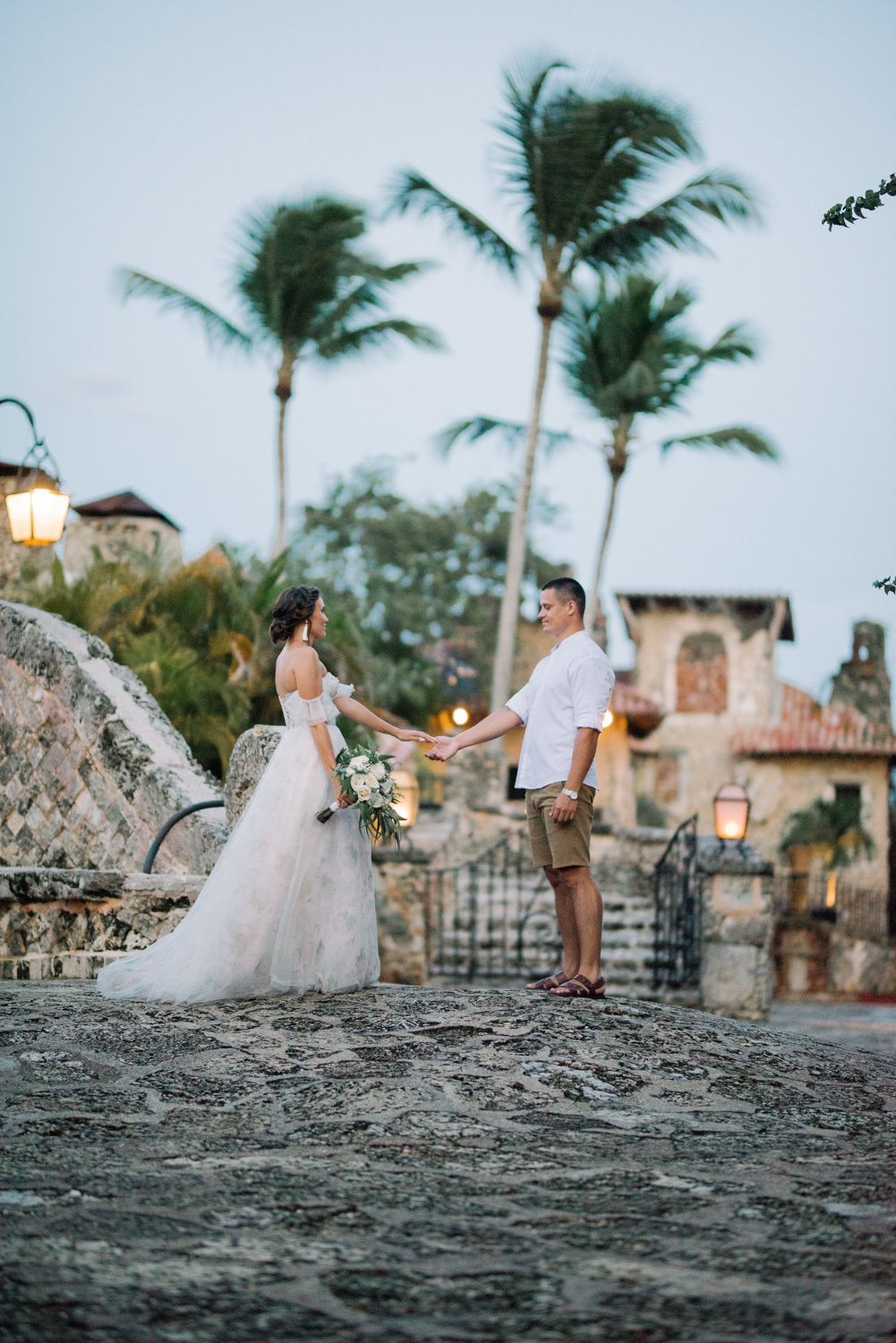 Жених и невеста держат друг друга за руку на каменном помосте перед каменным храмом на фоне трёх пальм.