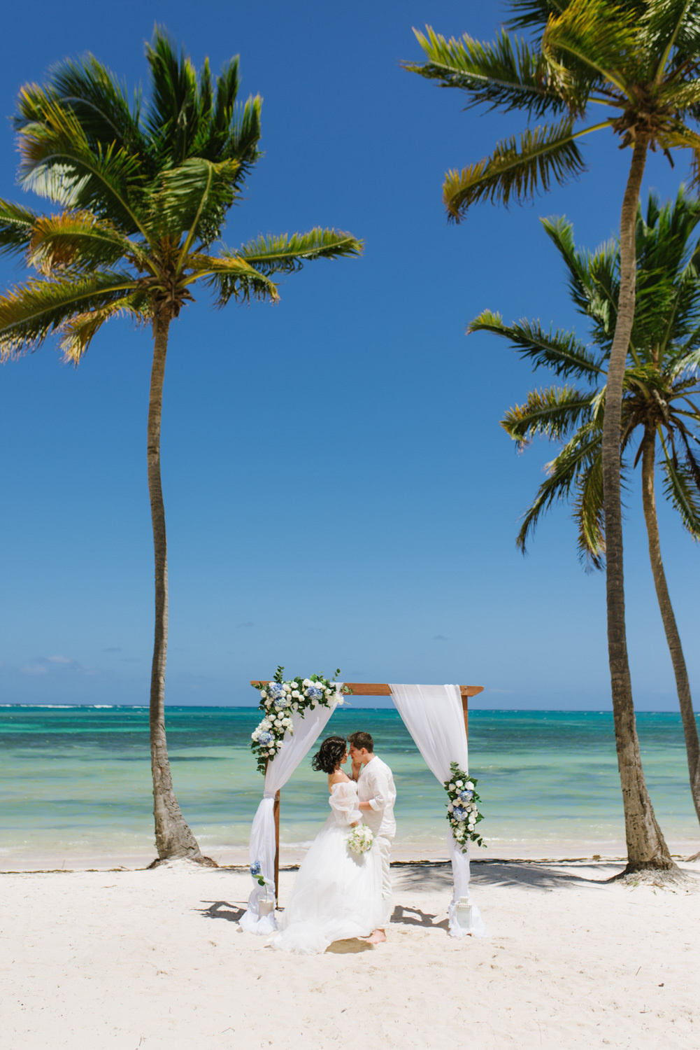 Bride and groom kiss on the beach with palm trees at the wedding altar on the background of the sea.