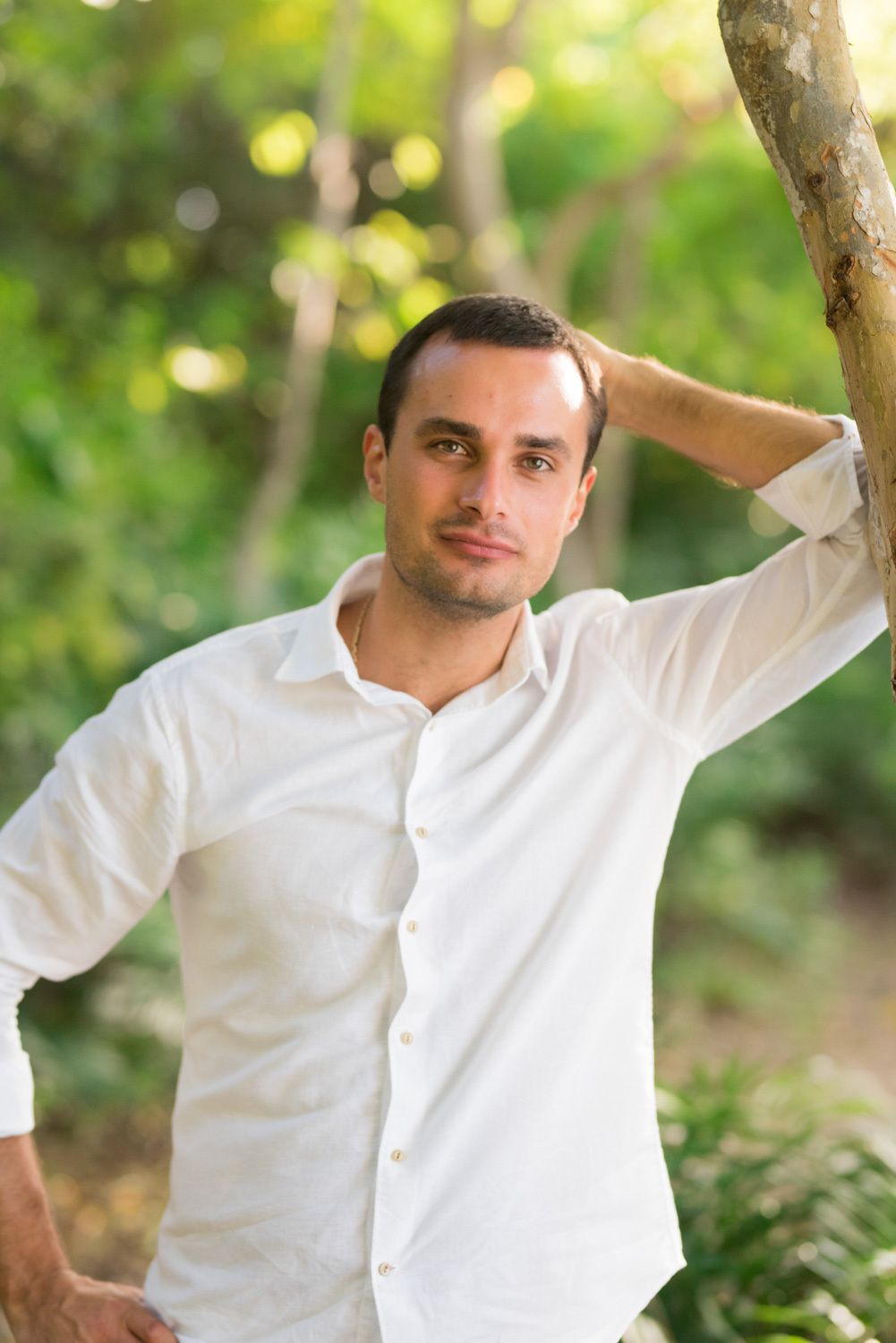 a man (groom) in a white shirt leans his elbow on a tree