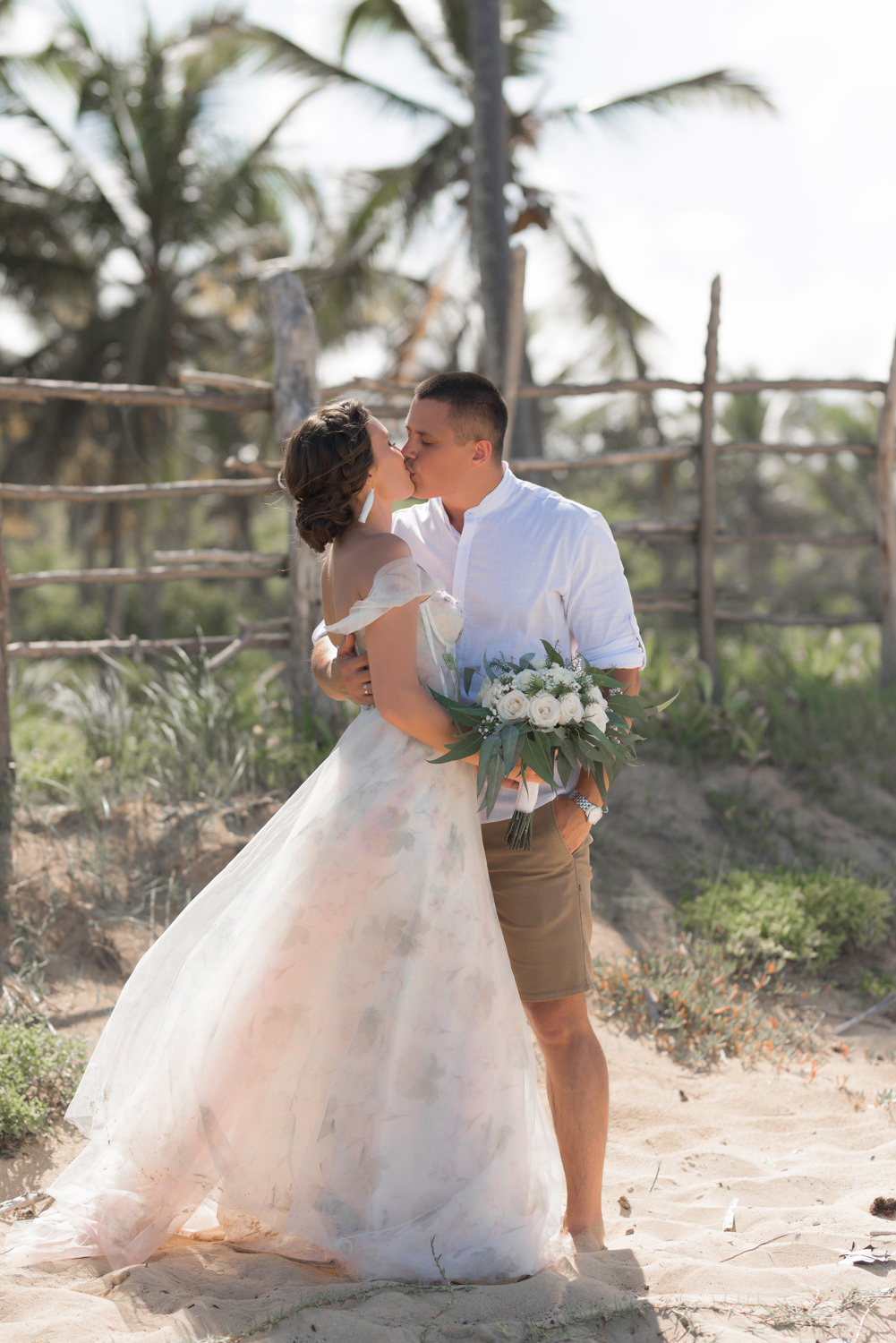 На берегу с пальмами у деревянного забора жених целует невесту.