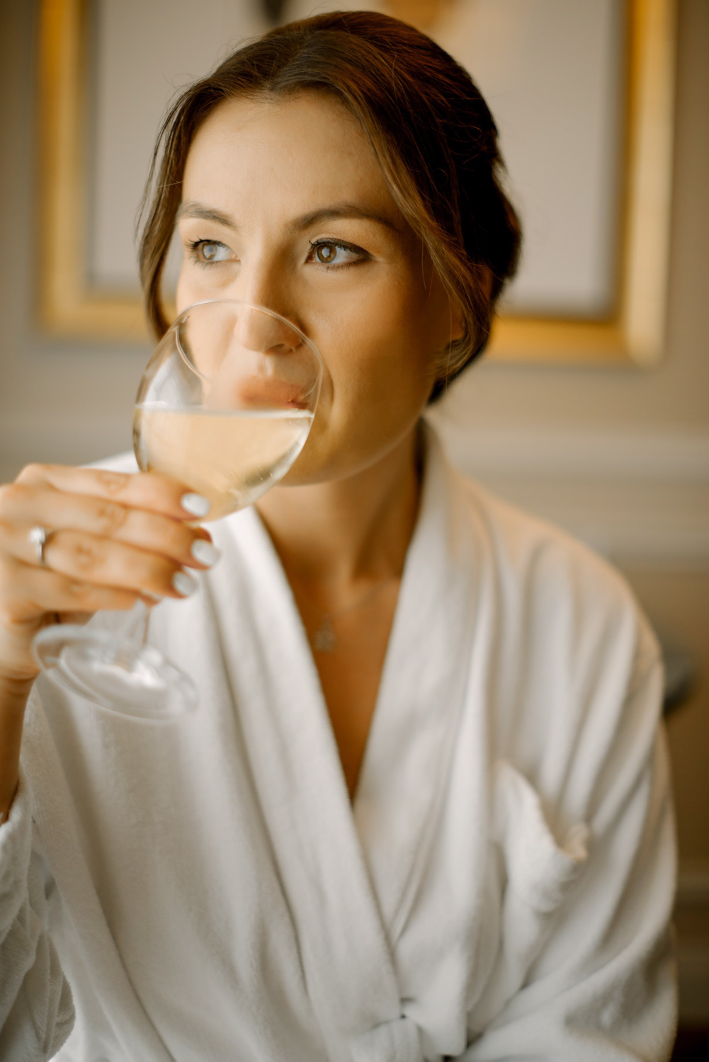 Девушка в домашнем белом халатике из бокала пьёт шампанское в своей комнате.