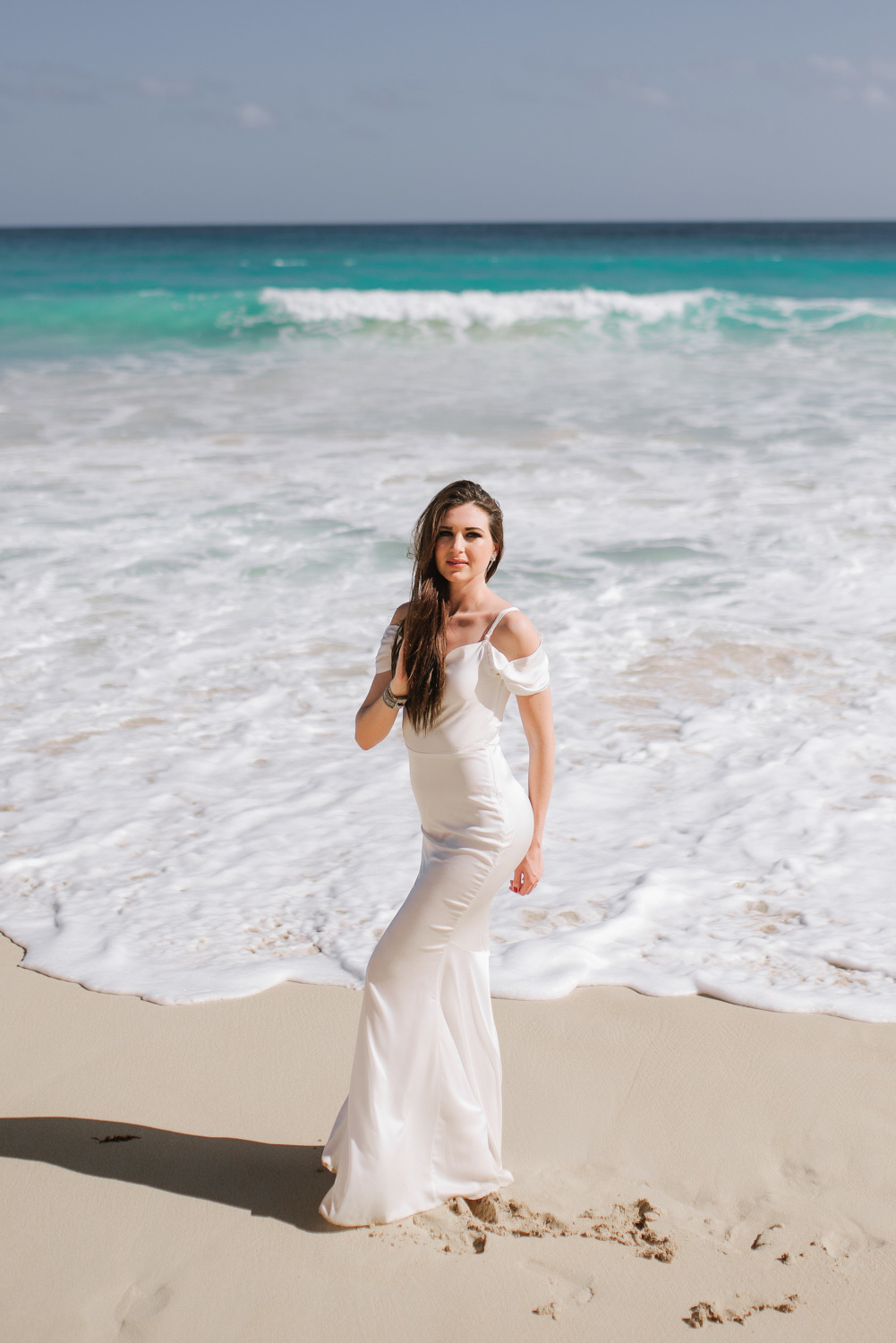 Bride in a white dress with long dark hair on the background of white foam waves and turquoise sea.