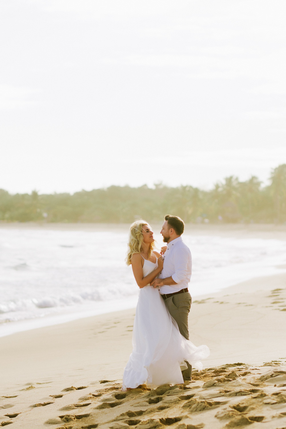 The bride in a white dress is hugged by the groom in a white shirt and gray trousers on the sandy beach against the background of the sea bay.