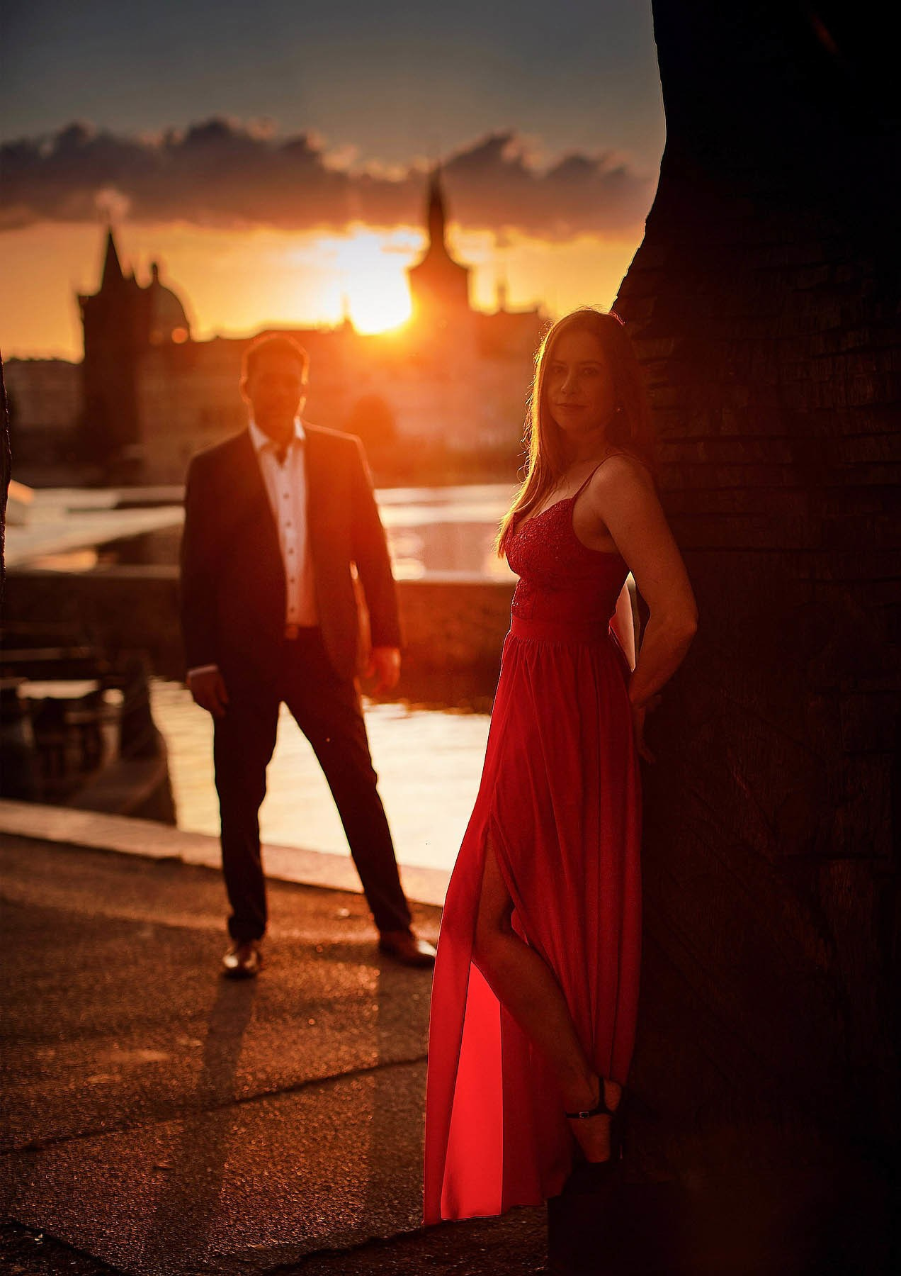 A woman wearing a red dress stands in a pose as her partner stands in the distance behind her as the early morning sun breaks above the skyline of Prague bathing them in a golden light
