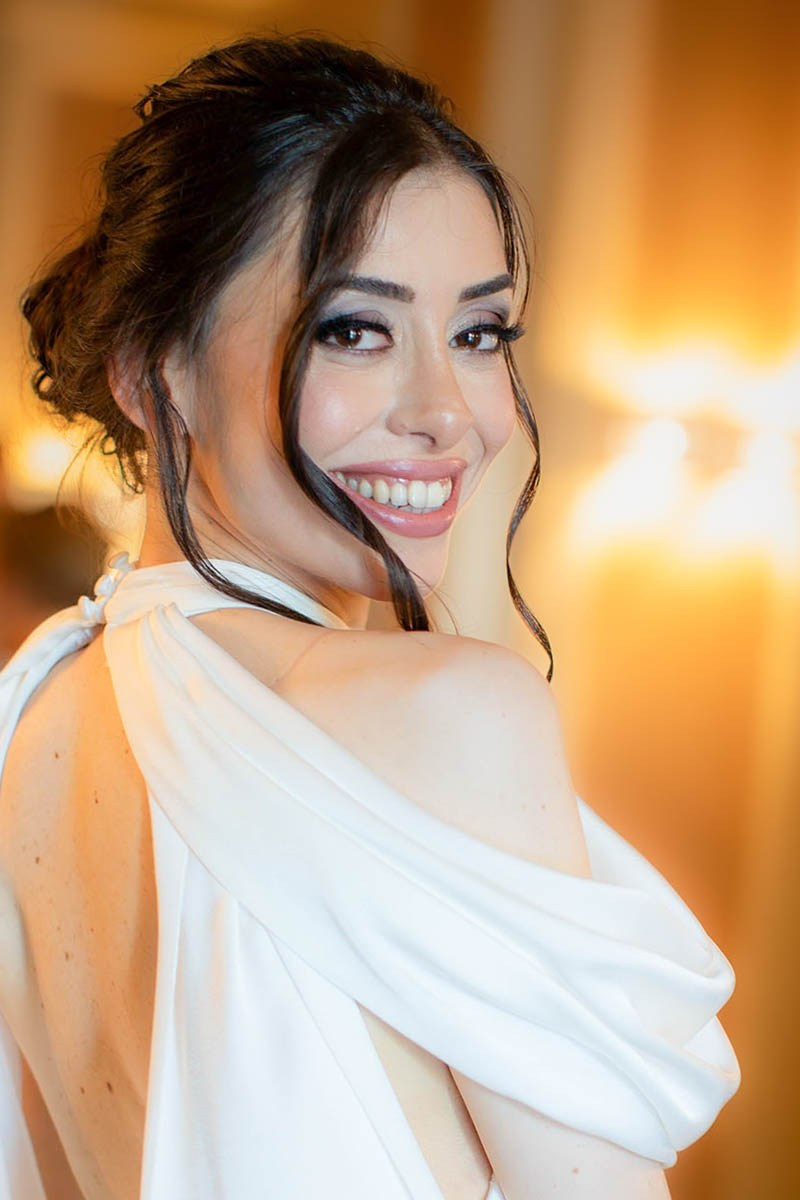 Radiant dark-haired bride looks over her shoulder as she smiles, surrounded by the warmth of indoor lighting.
