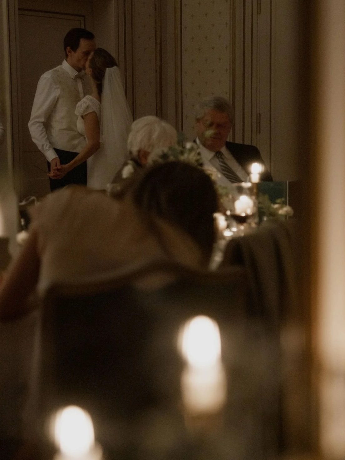 Couple sharing a quiet kiss in a candlelit ornate reception room, a flower-crowned guest seated in the warm foreground