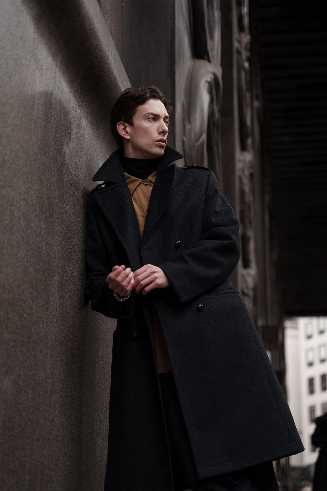 Male model in a long dark coat posing by a textured stone wall in Milan