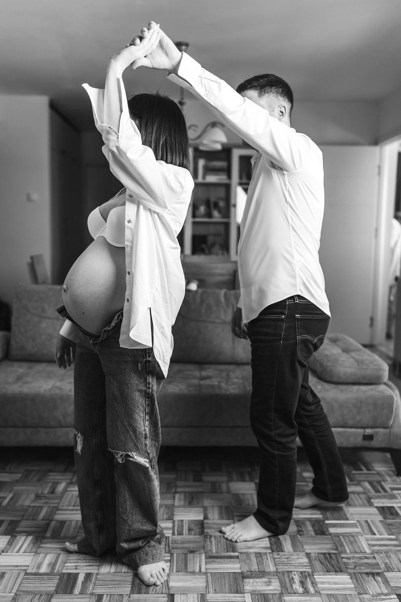 Crno-bela fotografija trudnice i partnera u dnevnoj sobi, uhvaćenih u spontanom trenutku plesa dok se drže za ruke.