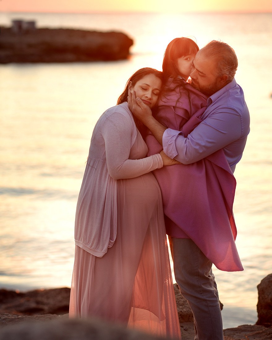 Family photoshoot at the seaside during sunset, parents and daughter embracing tenderly in warm light 