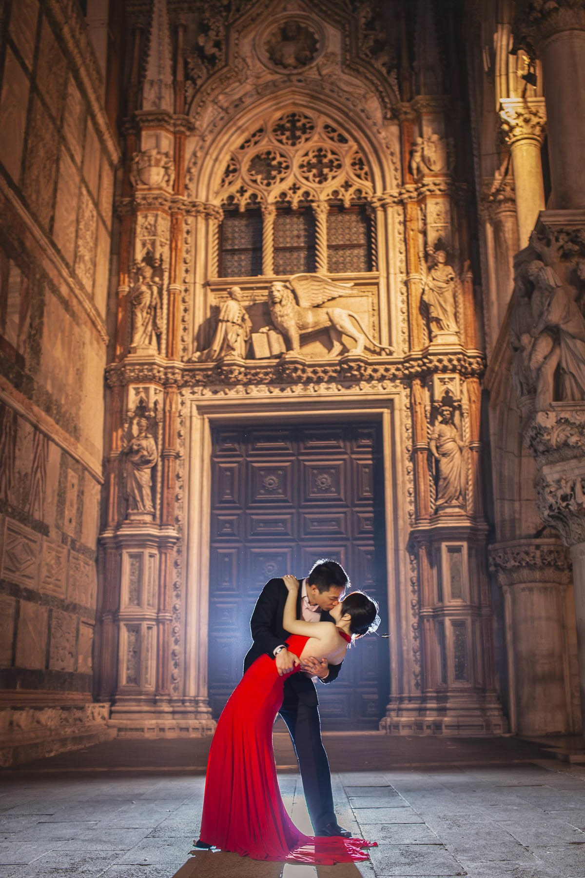 Groom dipping bride in red evening dress in front of Doge's Palace doors at night in Venice.