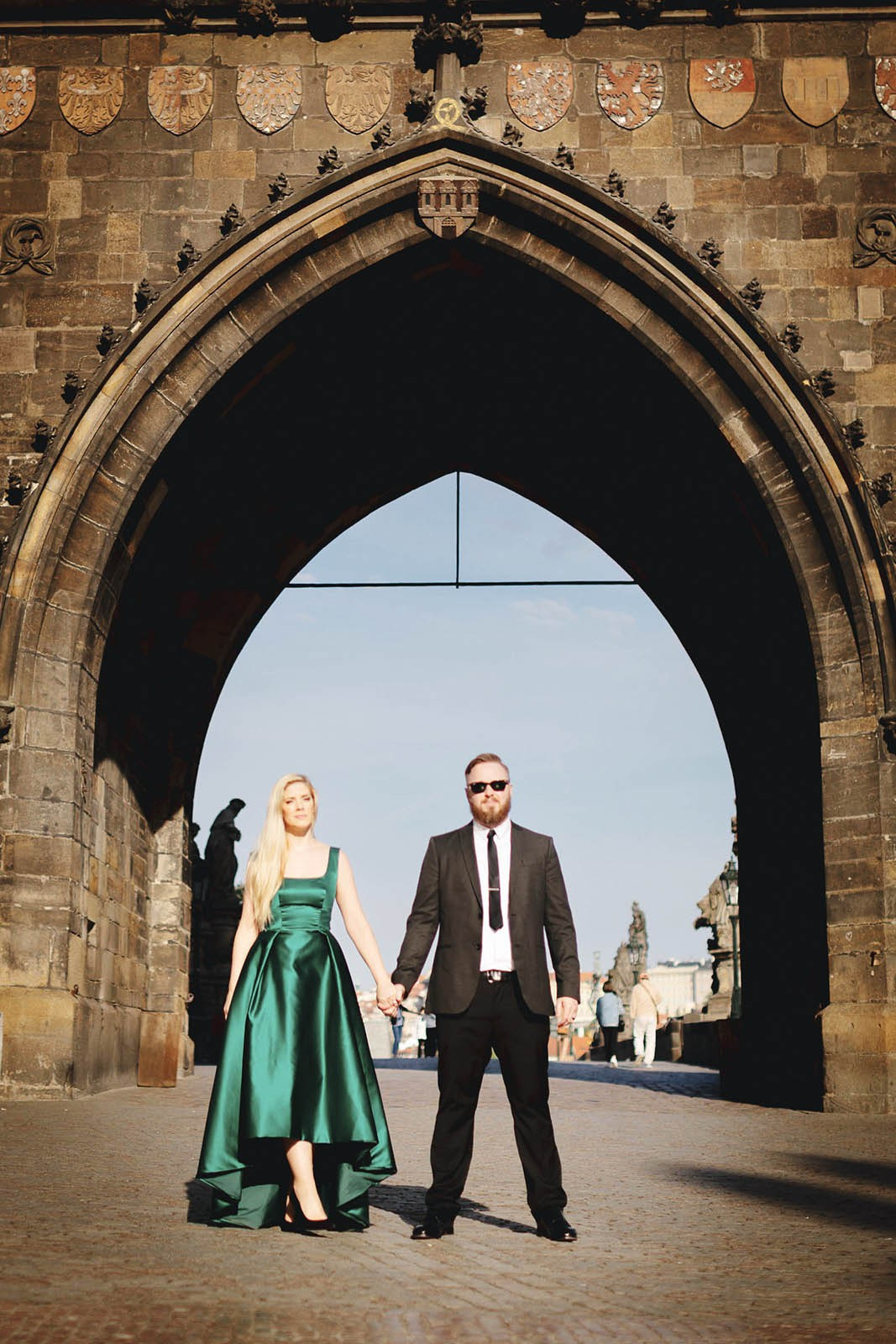 A woman in a green dress and a man in a suit stand under the Gothic tower near the Charles Bridge in Prague