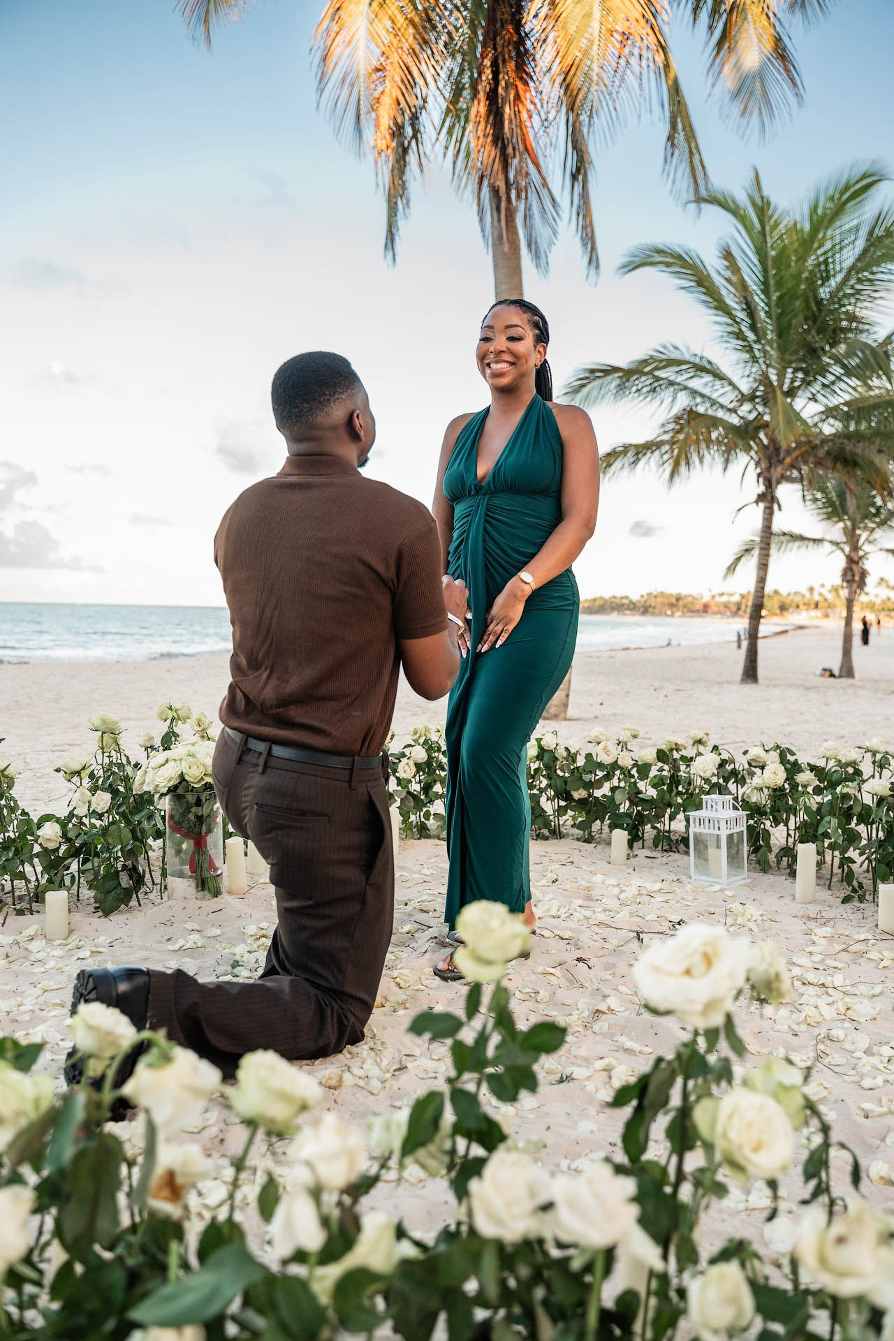 Man kneeling with an open engagement ring box as his partner reacts with excitement among fresh white roses.