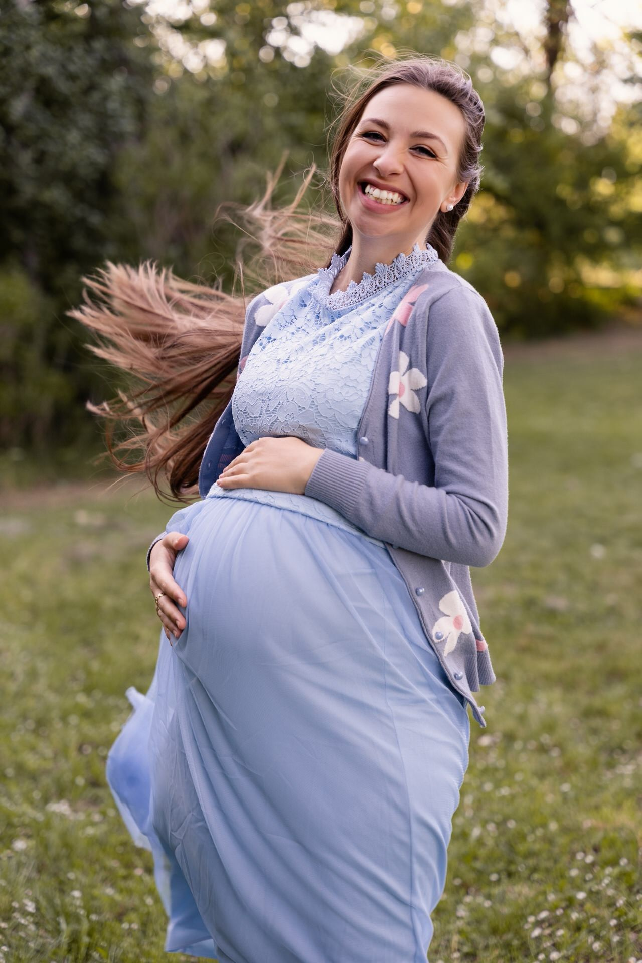 Nasmejana trudnica u svetloj haljini na travnjaku, okružena drvećem.