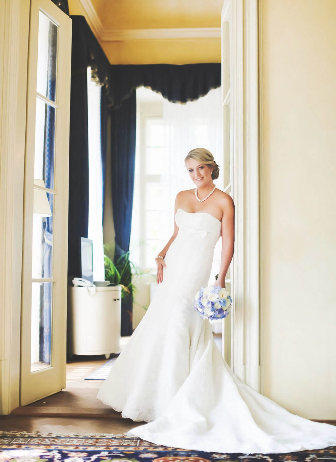 A smiling blonde-haired Czech bride leans against a wall while holding her bouquet before the start of her destination wedding at the Chateau Liblice.
