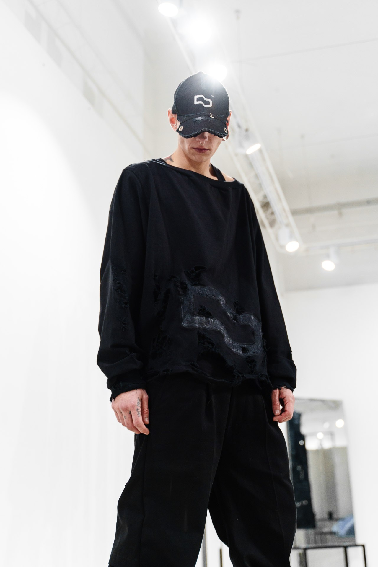 Edgy men's fashion portrait during Milan Fashion Week, featuring a model in a distressed black sweatshirt and a branded cap.