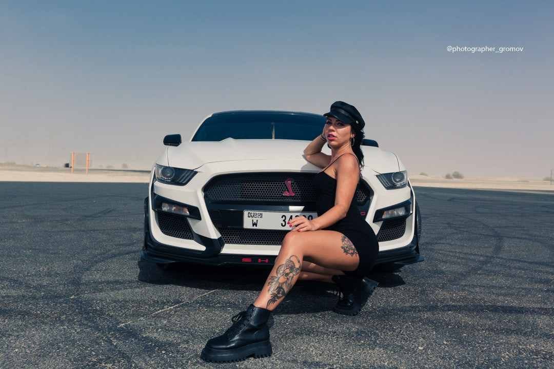 A girl in a black cap sits next to an exclusive, beautiful car against the backdrop of Dubai’s sandy roads
