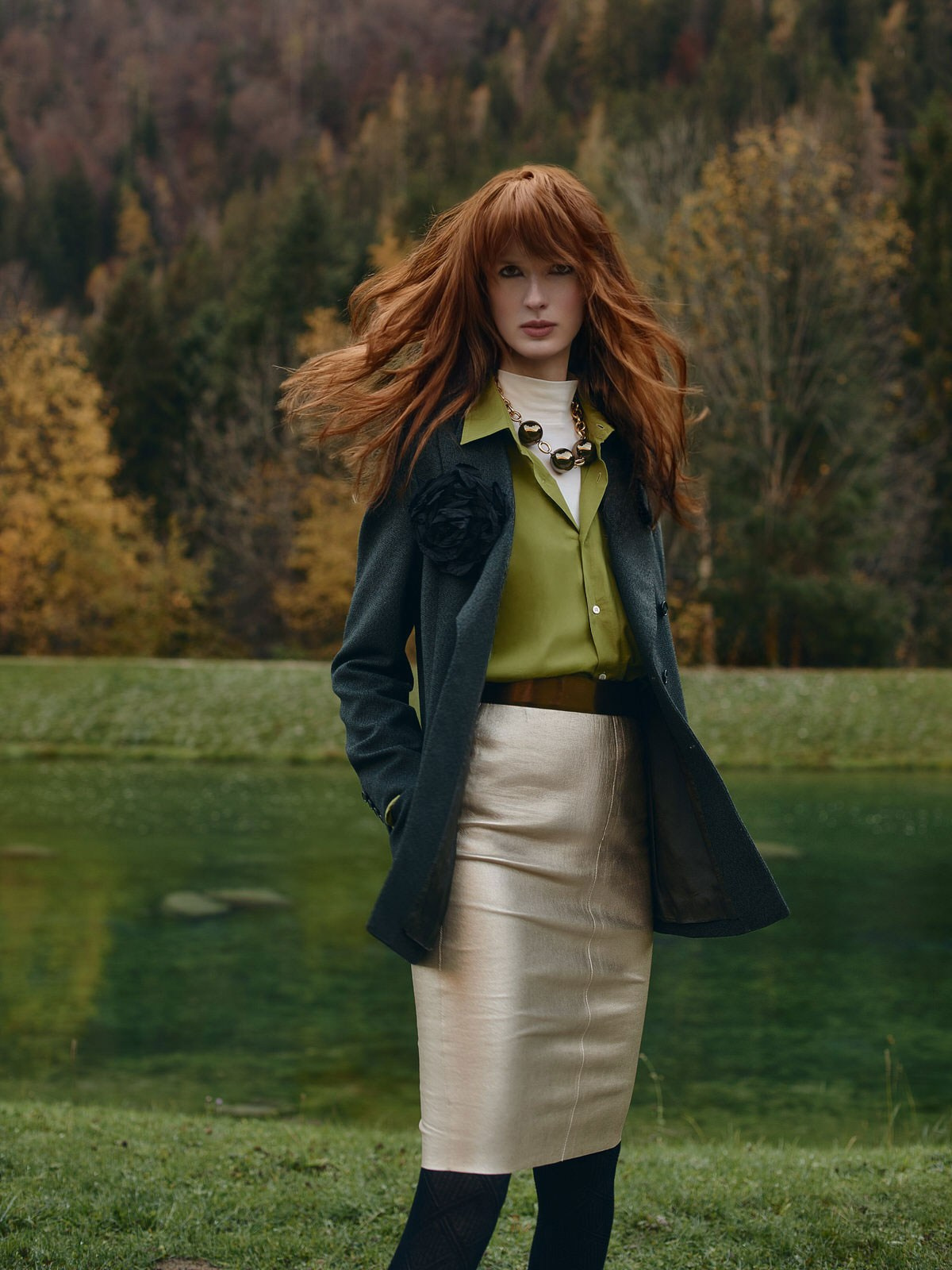 Elegant red-haired woman in green blouse and gold pencil skirt posing outdoors in autumn forest, fashion shoot in Italy