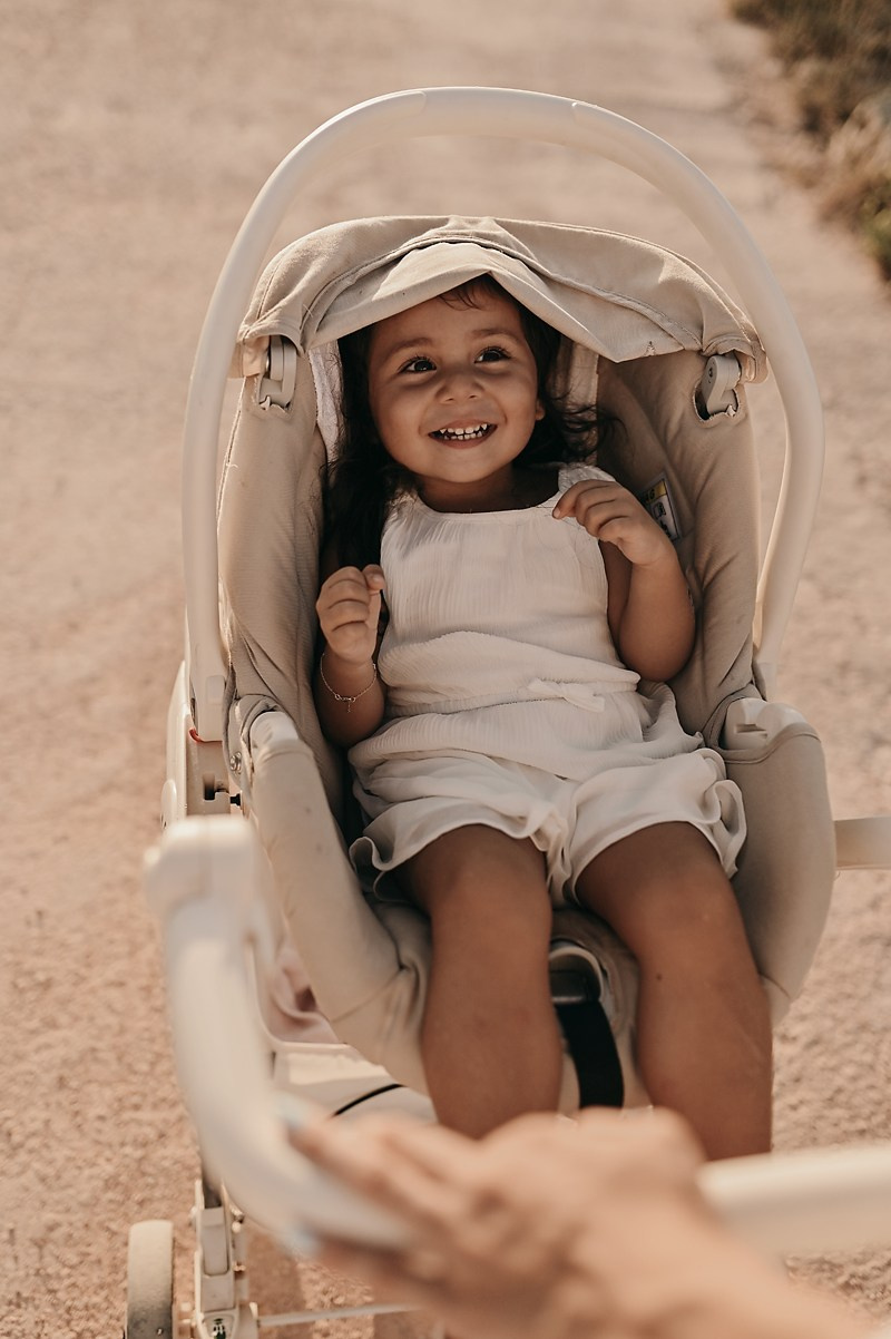 A seaside family walk at sunset in Puglia. A little girl laughs in her stroller as her mother pushes her along a sandy path.