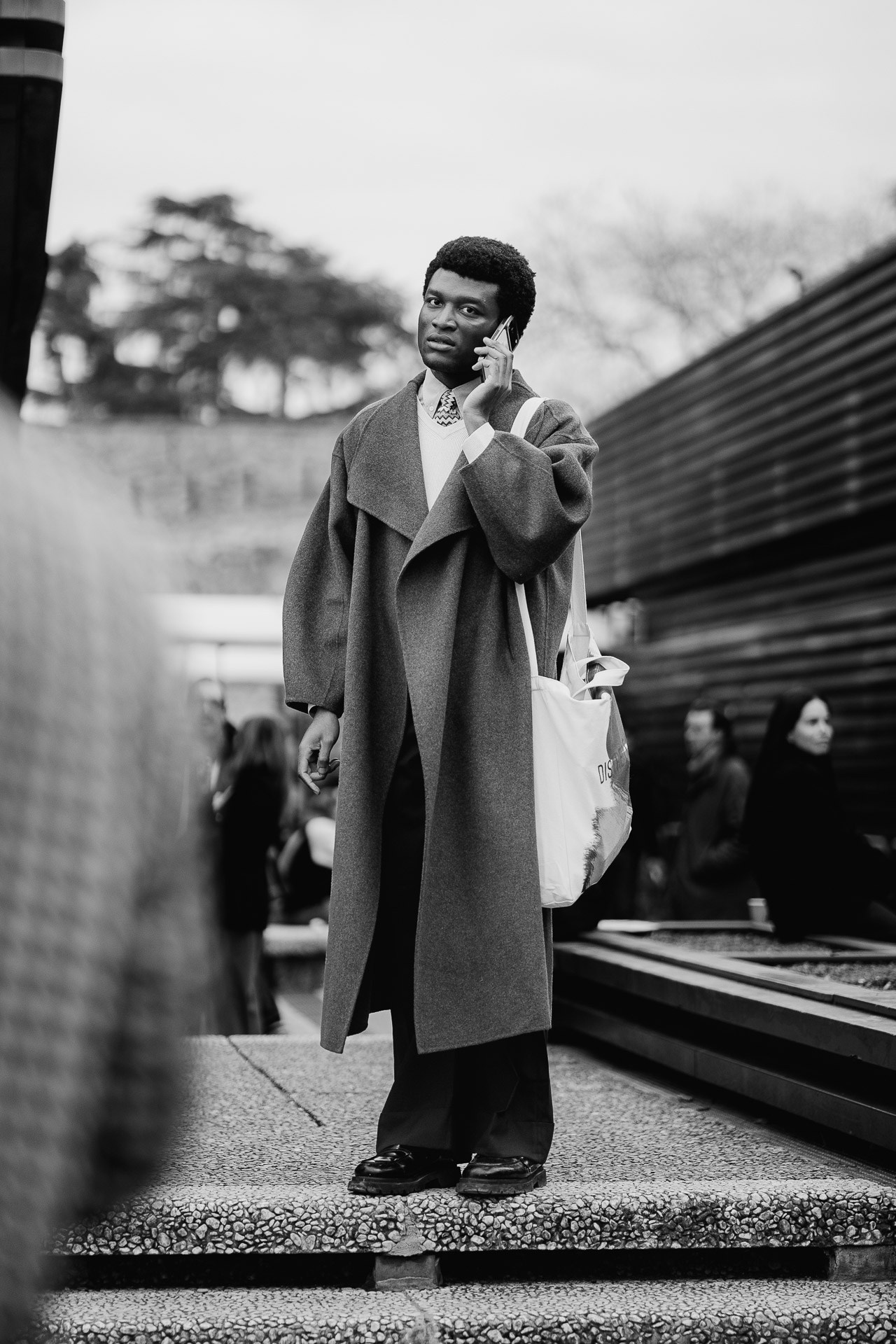 Black and white photo of man in long coat talking on phone at Pitti Uomo