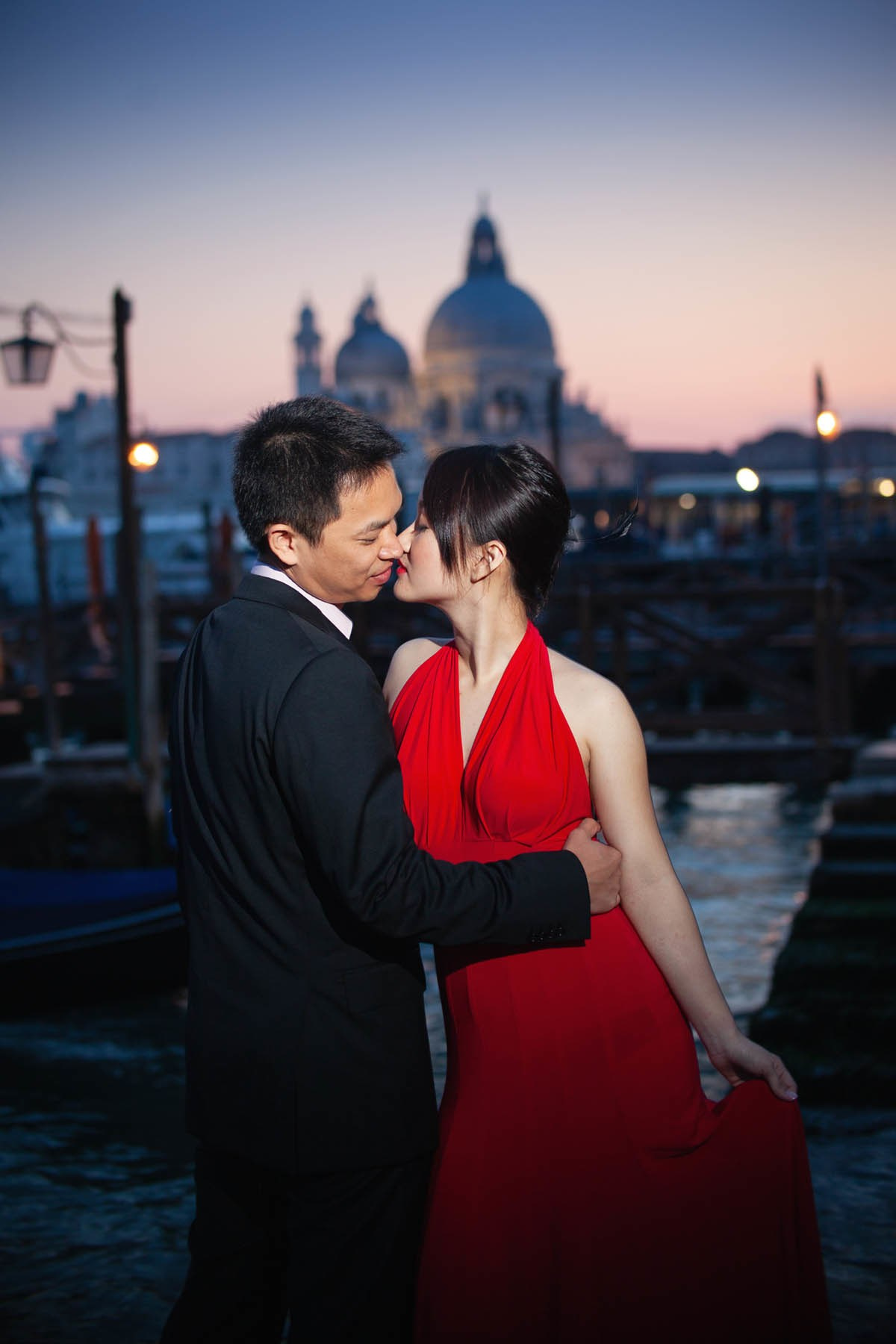 Thai bride in skin-tight red dress kissed by partner above gondola at sunset Venice.