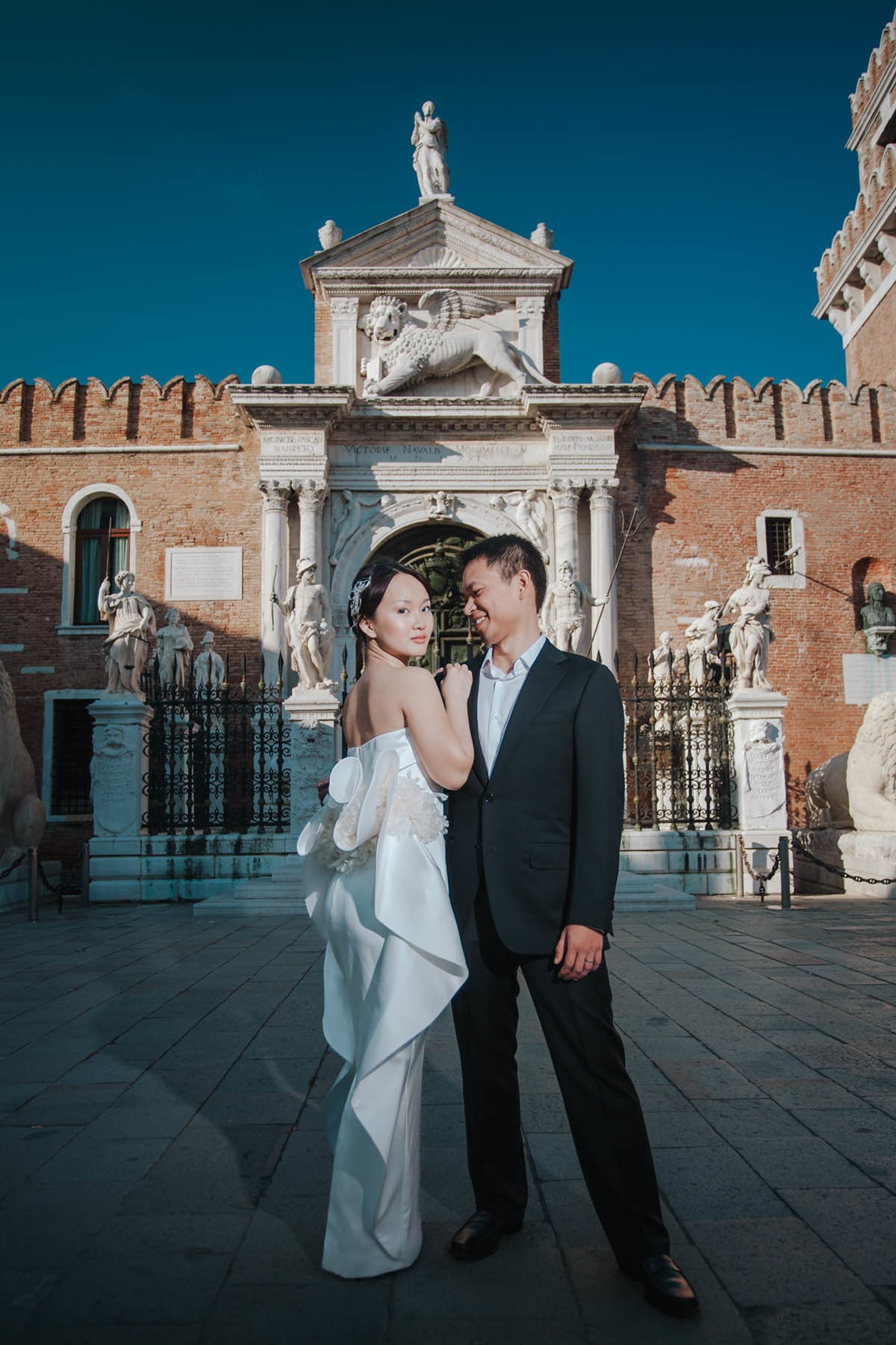 Smiling Thai bride embraced by groom in front of historic gate at Venetian Arsenal in Venice.