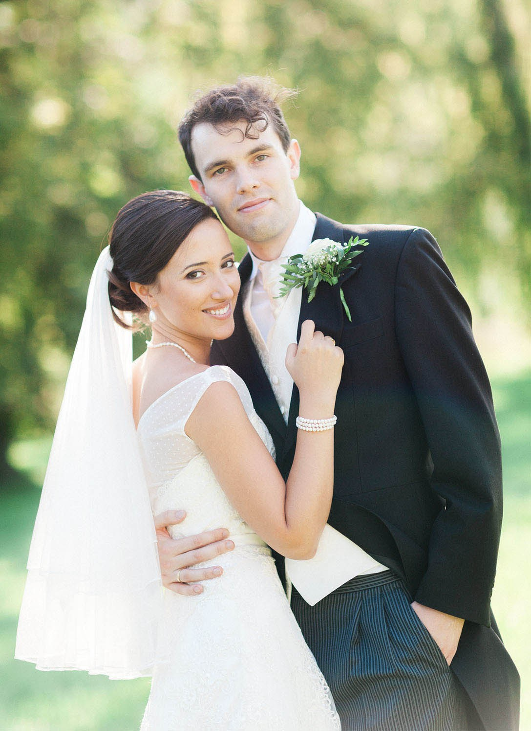 A smiling brunette bride and her tuxedo wearing groom smile at the camera during their wedding day portrait session in a garden setting.