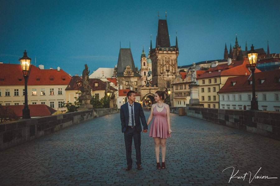 Woman in pink dress shocked as man proposes on knee with violinist performing on Charles Bridge at dawn.