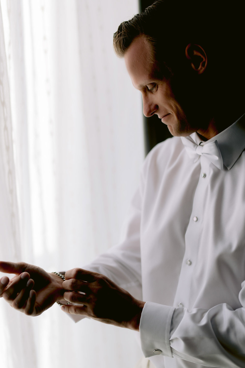 The groom puts on the bracelet while standing near the window in the hotel.