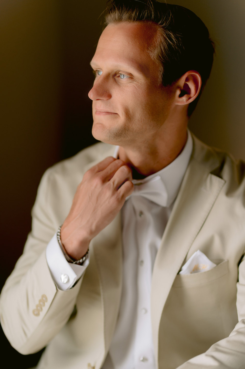 The groom is fixing his bowtie and looking out the window, he is wearing a light jacket and a white shirt.