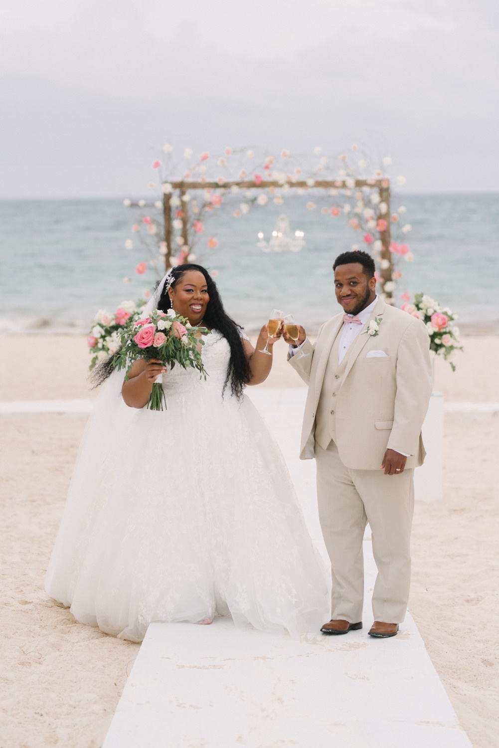 Жених и невеста позируют фотографу держась за руки. На заднем плане у них свадебная арка, украшенная розовыми цветами. 