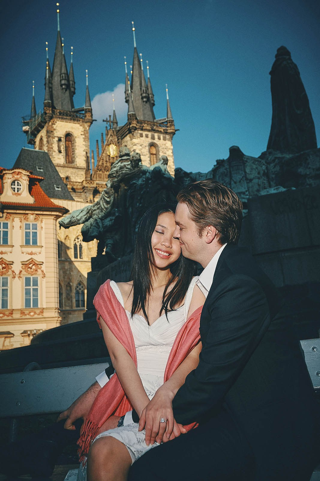 Couple sitting on a bench in Prague, enjoying the moment in the Old Town Square as Tyn Church towers above them