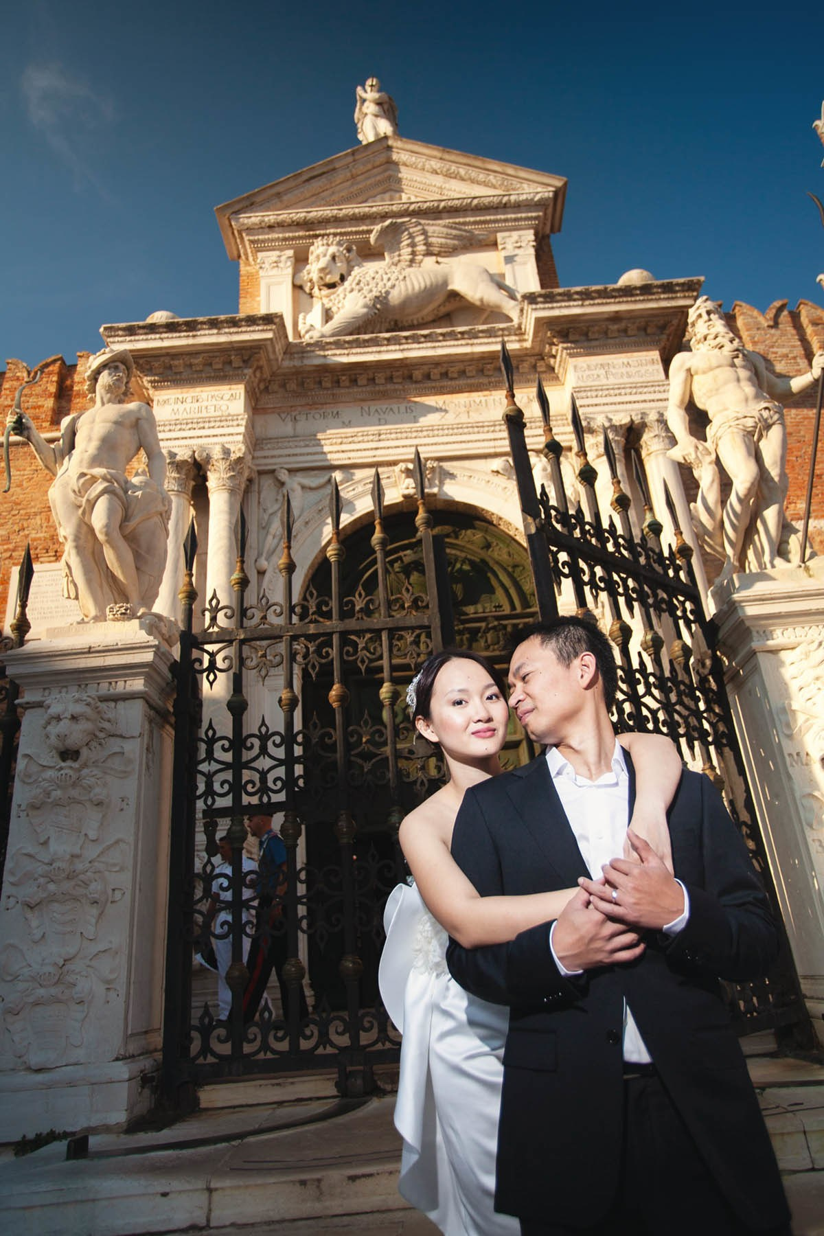 Groom smiling as bride embraces him from behind at Venetian Arsenal gate.