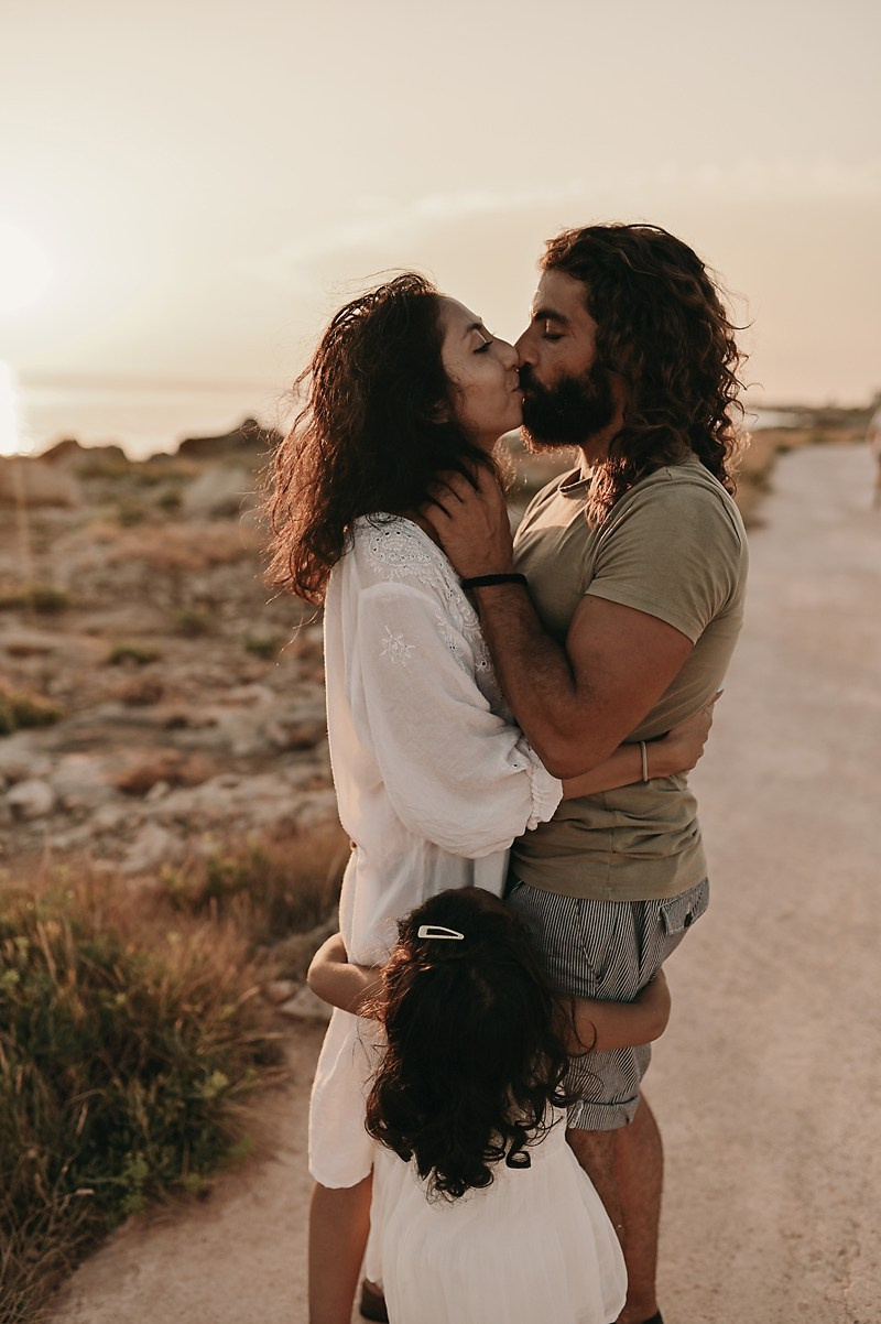 The parents share a kiss by the sea while their little daughter hugs them around the legs — a moment of deep family affection.
