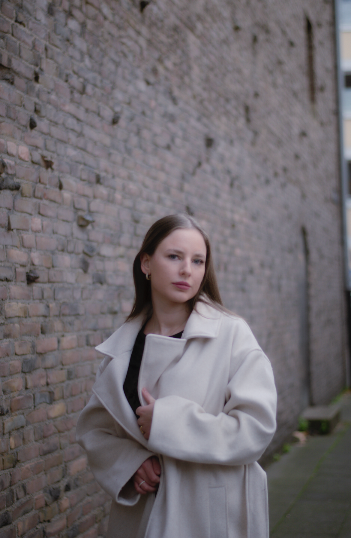 Porträt einer jungen Frau mit dunkelblonden Haaren, blauen Augen und hellbeigem Mantel vor einer braunen Mauerwerkswand