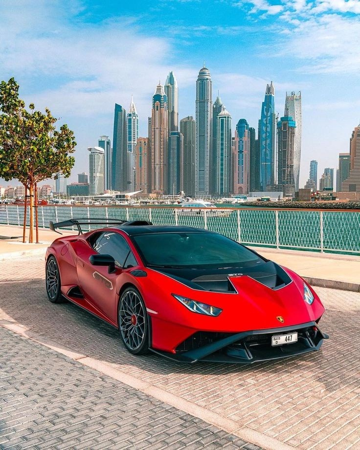 A red Lamborghini against the backdrop of Dubai Marina, parked on Palm Jumeirah. A sunny day with a clear blue sky.