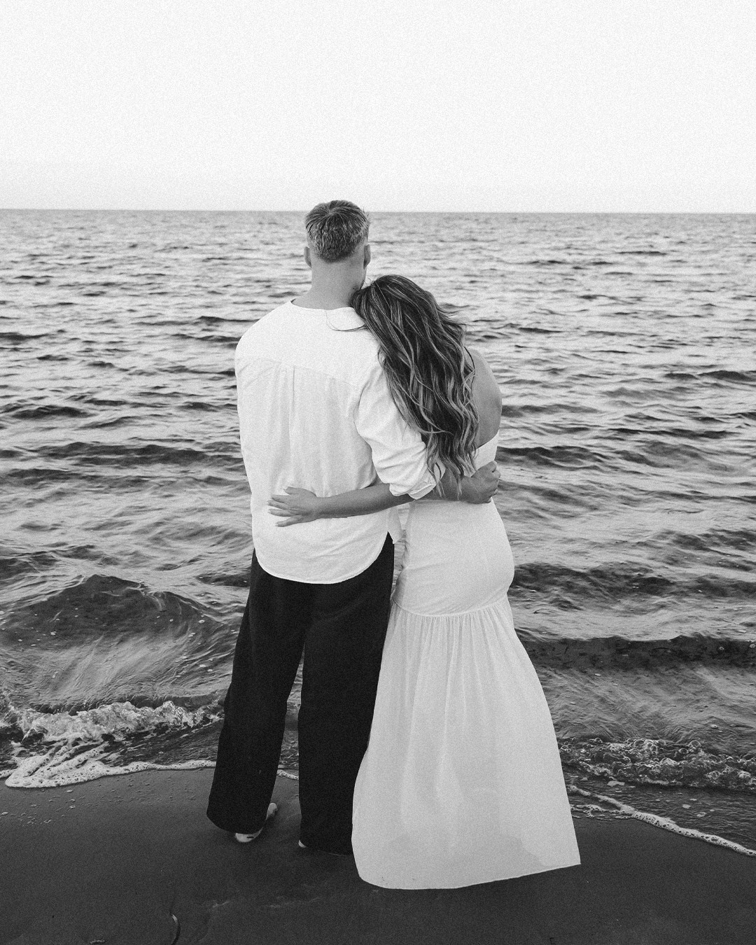 Pareja abrazada de espaldas frente al mar en blanco y negro.