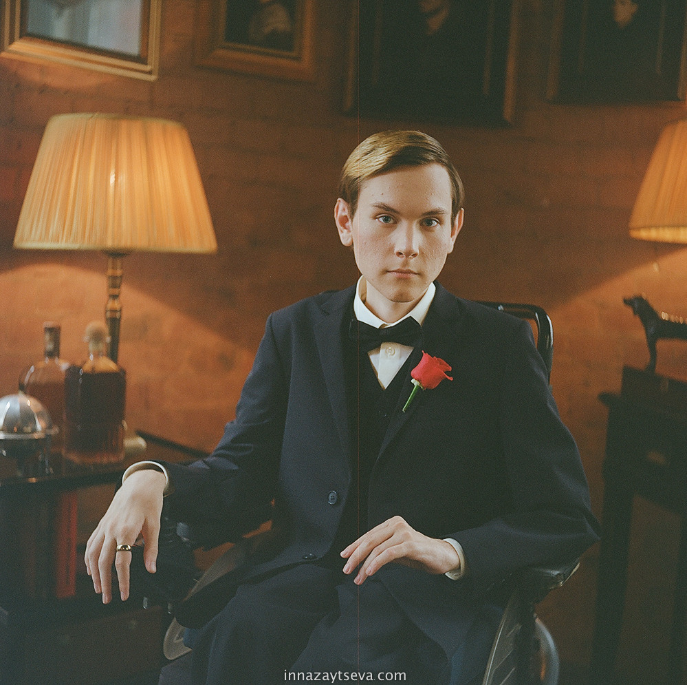 leistungsstarke Porträt junger Mann sitzt auf einem Rollstuhl in einer schwarzen Krawatte Anzug, Backstein Hintergrund mit warmen Lichtern, Porträt aus dem Wheeling Heart Projekt