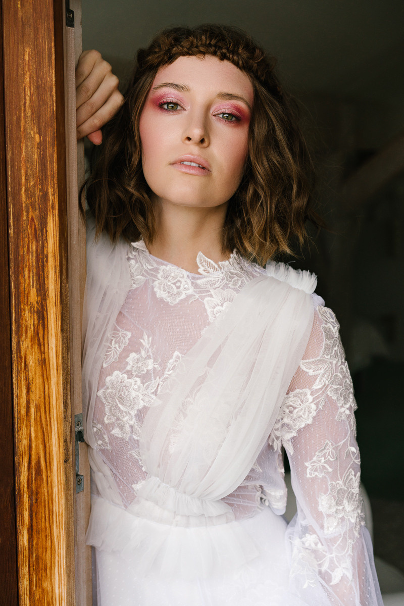 die junge Braut steht mit einer Hand im Türrahmen und trägt ein modisches rosa Make-up und ein Hochzeitskleid