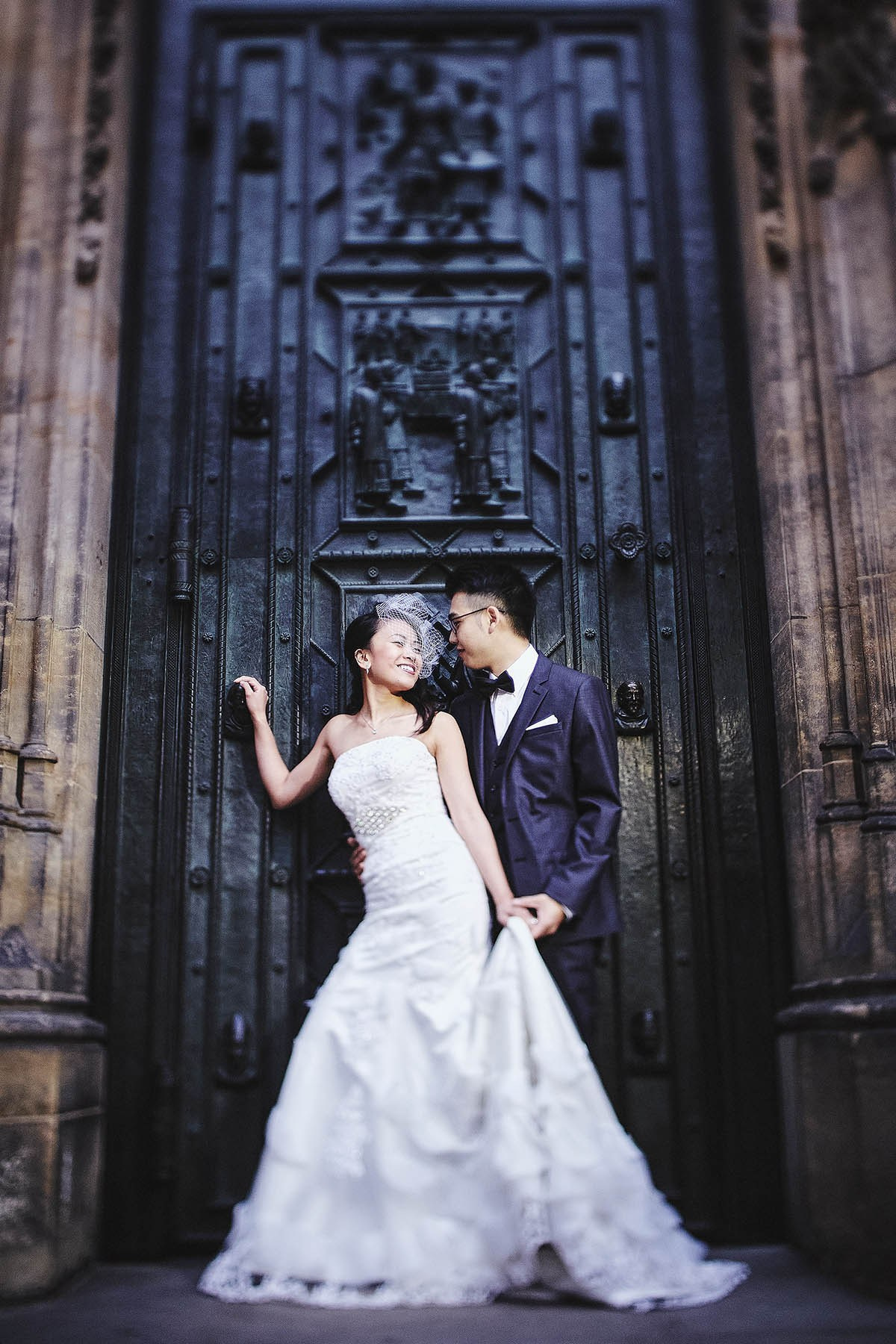 Bride in birdcage veil and vintage dress, embraced by tuxedo groom at St. Vitus Cathedral gates, Prague.