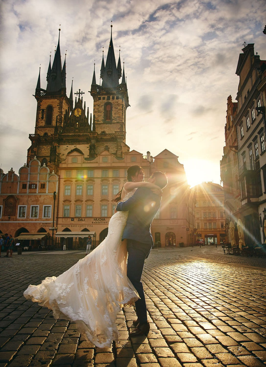 As the early morning sun breaks above the skyline of the Old Town Square in Prague, a young Asian bride is twirled by her groom in happiness