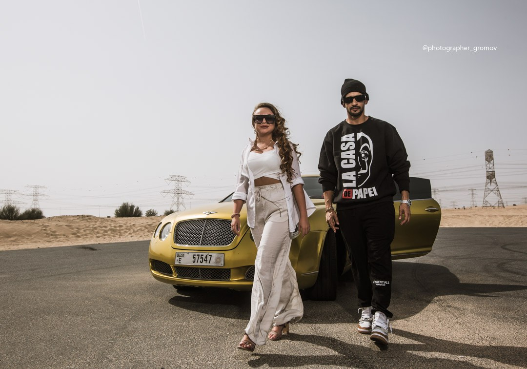 A young couple in sunglasses are walking near the luxury. Bentley cars against the backdrop of a Dubai sunset