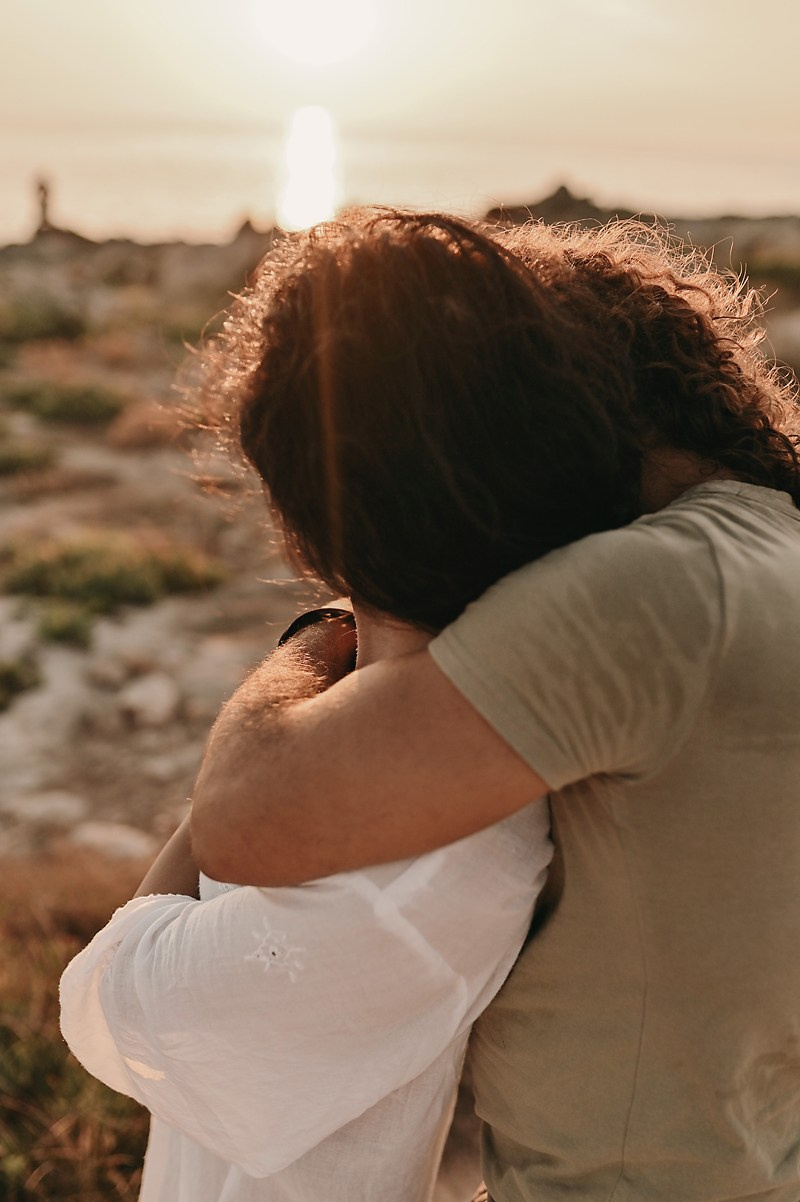 A couple embrace tightly with the sunset behind them, her face resting on his shoulder as the golden light shines through.