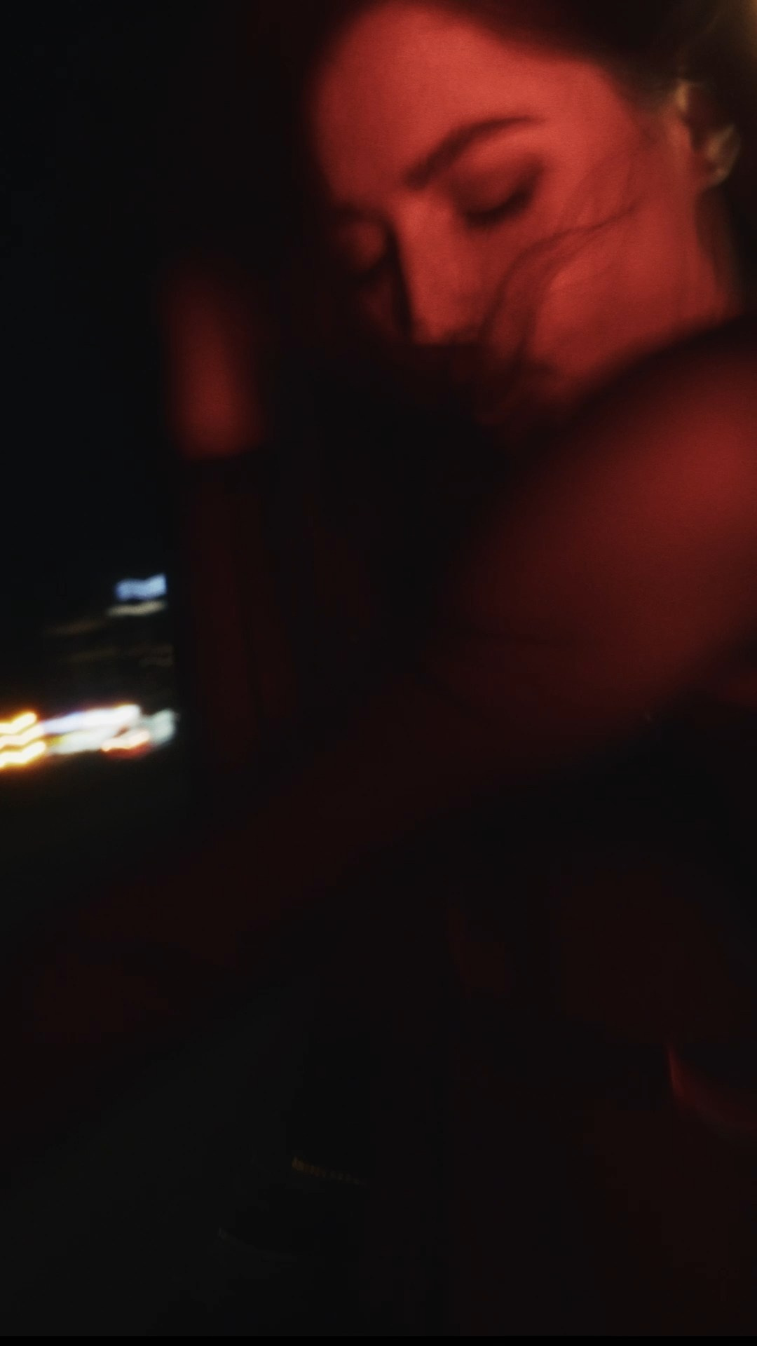 A softly blurred portrait of a person with closed eyes and flowing hair, illuminated by red and amber lighting against a dark urban backdrop.